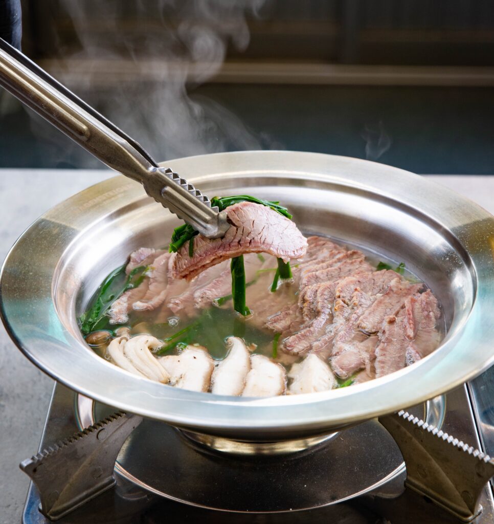 대구 북구 고성동 노원동 맛집 바름식당의 테이블 위 휴대용 버너에서 자작하게 끓여내어 김이 모락모락 나는 부드러운 1++ 못난이한우수육 한 점을 향긋한 부추와 함께 집게로 들어 올리는 순간의 상세 식감 연출 사진